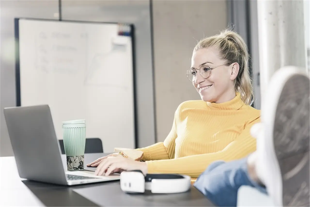 Smiling casual businesswoman sitting at table in office using laptop and eating a sandwich?